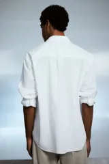 Camisa blanca de popelina de algodón con cuello inglés, tapeta clásica, dos bolsillos superiores abiertos, canesú con pliegue de caja en la espalda, mangas largas con puños abotonados, tapeta de manga con botón y bajo levemente redondeado. Corte holgado.