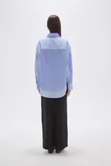 Camisa oversize de poliéster, con cuello camisero y cierre frontal de botones. Presenta un diseño de rayas verticales azules y blancas, con bloques de rayas horizontales en los puños y el ruedo inferior.