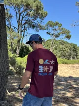 Remera de manga corta color bordó con un estampado gráfico en la espalda que incluye un sol amarillo, una ola azul, una estrella de mar blanca, un símbolo estilizado en azul y la frase "VITAMIN SEA" en azul.