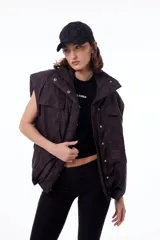 Chaleco negro con estampado de logos, cuello alto, cierre frontal con cremallera y broches a presión ocultos, bolsillos delanteros y cintura ajustable con cordón elástico. Interior de tafeta relleno de guata.