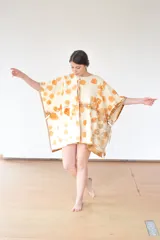 Capa de lana color crudo con estampado de hojas de eucalipto en tonos naranja y marrón. Tiene cierre con broches a presión en el escote y ojales metálicos con cintas para anudar en los laterales.