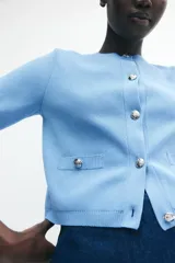 Cárdigan corto de punto fino celeste, con cuello redondo, manga corta y cierre frontal con botones metálicos decorativos.