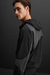 Cazadora con capucha confeccionada en tejido técnico ligero y elástico en todas las direcciones, color negro con detalles en gris. Cuenta con cuello con capucha ajustable, manga larga, bolsillos con cierre de cremallera en cadera, acabados con elástico y cierre frontal de cremallera.
