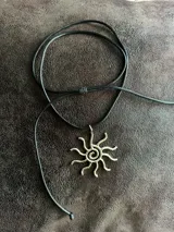 Collar con cordón negro ajustable y dije dorado en forma de sol con espiral en el centro.