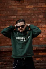 Buzo verde oscuro de algodón con cierre medio frontal, cuello alto y corte oversize. Estampado frontal con la frase "Against All" en color crema y detalles bordados.