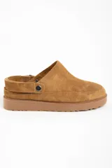 Zueco tipo mule de gamuza color camel, con forro interior de corderito visible en el borde. Presenta una tira ajustable con broche metálico en el lateral y suela de goma gruesa y dentada.