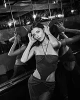 Vestido largo negro, ajustado al cuerpo, con escote strapless y tirantes finos cruzados en el cuello. Presenta recortes laterales pronunciados en la cintura y un nudo fruncido en la parte inferior frontal.