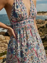 Vestido largo con estampado floral y de aves en tonos rojizos y azules sobre fondo crema. Presenta escote halter profundo en V y cintura marcada con falda con vuelo en capas.