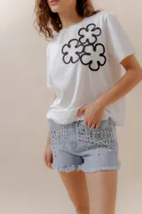Remera blanca de algodón con cuello redondo y manga corta. Presenta un aplique frontal con tres flores estilizadas bordadas en hilo negro.
