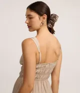 Vestido corto color beige, con escote recto, breteles medianos y lazo decorativo en el centro del busto. Presenta costura fruncida en la cintura y falda con babados en capas.