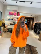 Buzo tipo canguro oversize de tela plush, color naranja vibrante, con capucha y cordones de ajuste en color rosado pálido. Presenta mangas largas con puños fruncidos y un bolsillo canguro frontal.