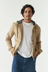 Campera de felpa color beige con capucha y cordón ajustable, cierre frontal, bolsillos con cierre, lettering estampado en la espalda y logo en grifa de goma.