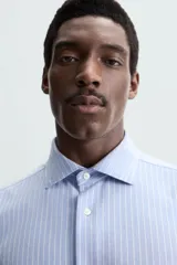 Camisa celeste a rayas verticales blancas, de corte recto, con cuello italiano abierto, mangas largas con puños abotonados y cierre frontal con botones.