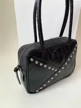 Cartera rígida negra de cuero con tachas plateadas y tres combinaciones de cuero diferentes que aportan textura. Tiene herrajes y cierre metálico plateado marca YKK. Cuenta con un asa fina para llevar al hombro o en la mano y una correa larga extra regulable.