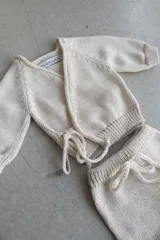 Conjunto tejido artesanal en hilado color crudo, compuesto por un cárdigan cruzado con lazos y un pantalón con cintura ajustable con lazos.