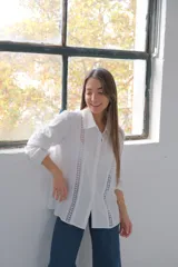 Camisa blanca de manga larga con puños, cuello camisero y cierre frontal con botones ocultos. Detalles de entredós a tono.
