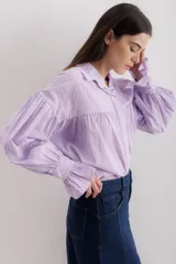 Camisa gris a rayas verticales blancas, de corte amplio, cuello alto con lazo y mangas largas abullonadas con puños elásticos.