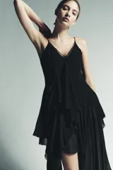 Vestido midi negro, con escote pico adornado con encaje a tono y tirantes finos. Presenta una cascada de volantes en la parte delantera y un bajo asimétrico.