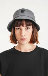 Gorro tipo bucket de color gris oscuro (carbón), con ala ancha y un pequeño parche negro con un logo bordado en blanco en el frente.