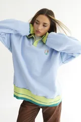 Buzo celeste con cuello polo y puños en rib color verde lima. Tiene un parche con el logo de la marca en el pecho y rayas horizontales en el ruedo.