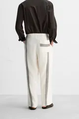 Pantalón relaxed fit confeccionado en tejido de lino color crudo. Cintura elástica ajustable con cordón, bolsillos laterales y traseros. Perneras con aplicación de bandas verticales negras a contraste en delantero.