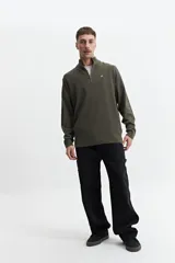 Sweater verde oliva de punto con cuello alto y cierre de medio cierre. Tiene puños y logo bordado en el pecho.