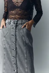 Falda midi de denim rígido, color gris claro lavado, con botones metálicos grabados en el frente y recortes verticales.