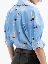 Camisa de poplin satinado color celeste con estampado de cerezas y rayos amarillos. Presenta mangas tipo bombé con puño y cierre frontal con botones ocultos.