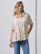 Blusa color crema de crepe con cuello a la base con solapa, escote en V con abertura central y botones. Delantera con detalles plisados y apliques en crochet. Manga corta con terminación fruncida y ruedo en broderie.