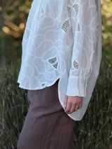 Camisa blanca de broderie con diseño floral, corte oversize, cuello clásico, cierre frontal con botones y mangas largas con puños abotonados.