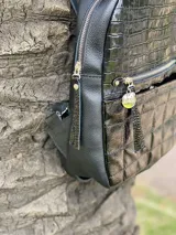 Mochila pequeña negra con textura tipo cuero de cocodrilo en la parte superior y diseño acolchado en la parte inferior. Tiene un bolsillo frontal con cierre, correas ajustables y asa superior.