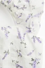 Blusa blanca con estampado floral en tonos lavanda, de tela ligera, con escote recto, breteles anchos con volados, espalda escotada con tiras finas horizontales que se atan en la parte superior, sección de punto smock en la espalda baja, costura fruncida en la cintura y peplum con vuelo.