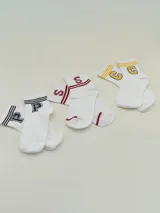 Pack de tres pares de medias tobilleras blancas con detalles de letras bordadas en contraste: un par con letras 'P' en negro, un par con letras 'S' en rojo y un par con letras 'C' y 'G' en amarillo y negro. Todas tienen detalles de rayas en el borde superior.