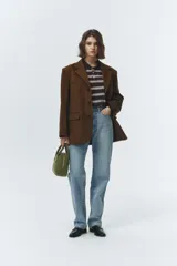 Chaqueta de gamuza marrón con diseño sastrero y corte levemente oversized. Presenta solapas de muesca, cierre frontal con botones y bolsillos laterales con solapa. Confeccionada en cuero vacuno con acabado tipo suede y forro de poliéster.