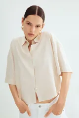 Camisa color crema en tejido liviano de lino y viscosa con cuello, botones adelante y pliegue de caja invertido en la espalda. Modelo de corte holgado con hombros marcadamente caídos, mangas al codo y bajo levemente redondeado.