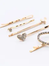 Set de cinco hebillas doradas para el pelo, con diferentes diseños: una con forma de corazón con strass, una con forma de corazón liso, una con strass, una lisa y una retorcida.