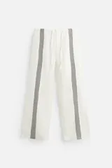 Pantalón relaxed fit confeccionado en tejido de lino color crudo. Cintura elástica ajustable con cordón, bolsillos laterales y traseros. Perneras con aplicación de bandas verticales negras a contraste en delantero.
