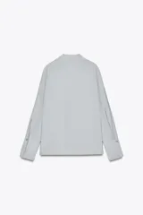 Camisa celeste fluida confeccionada en hilatura con mezcla de lyocell. Cuello redondo, manga larga con puños y bajo asimétrico con aberturas laterales. Cierre frontal con botones.