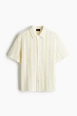 Camisa de manga corta color crema, confeccionada en tejido de punto jacquard con textura calada. Tiene cuello inglés, tapeta clásica con botones y bajo recto.