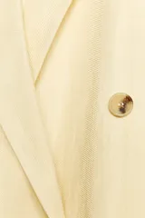 Blazer color crema confeccionado en hilatura con lino, de corte recto, con cuello solapa de muescas y manga larga acabada en abertura y botones. Tiene bolsillos delanteros y cierre frontal cruzado con botones.