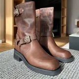 Bota de cuero vacuno color marrón, con suela de goma artesanal, caña ajustable con hebilla y cierre interno YKK.