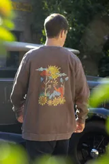 Buzo marrón con efecto lavado, cuello redondo y mangas largas. Estampado en la espalda con un diseño de estilo cartoon que incluye un sol, nubes, hongos y flores.