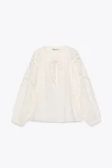 Camisa color crudo confeccionada en mezcla de algodón y lino, con cuello redondo y escote pico ajustable con lazadas. Tiene manga larga abullonada y detalle de bordados perforados a tono.