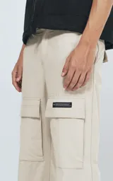 Pantalón cargo de color beige claro, corte recto y holgado, con bolsillos tipo parche en los muslos. Presenta costuras reforzadas en las rodillas y una etiqueta rectangular negra en el bolsillo derecho.