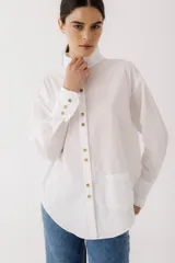 Camisa blanca de poplin con cuello alto con volados, cierre frontal con botones dorados, manga larga con puños con volados y bolsillo de parche en el frente.