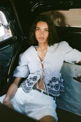 Blusa de rayas verticales azules y blancas, con escote en pico, mangas cortas abullonadas con elástico y bajo con bordados florales perforados. Cierre frontal con lazos.