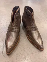 Botín corto de cuero color plata metalizado, con punta fina y cierre lateral. Presenta tachas metálicas decorativas en el empeine y en una tira que rodea el tobillo. Tiene un taco bajo y suela de color natural.