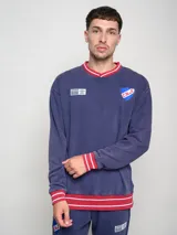 Buzo azul marino de felpa con efecto lavado, cuello en V y puños en color rojo con rayas blancas. Presenta el escudo del Club Nacional de Football y el logo de Umbro bordados en el pecho.