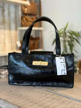 Cartera tipo tote bag baguette de cuero charolado color negro, con cierre dorado y doble asa corta.