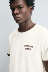 Camiseta color crudo de corte relaxed fit, con cuello redondo y mangas cortas con ribete marrón. Estampado frontal con la frase "Golden Hour" en color marrón.